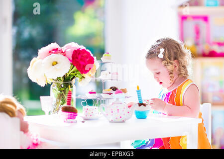 Adorable toddler girl with curly hair wearing a colorful dress on her birthday playing tea party with a doll, toy dishes Stock Photo