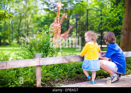 Happy school boy and his toddler sister cute little girl with curly hair wearing a dress in a zoo watching giraffes Stock Photo