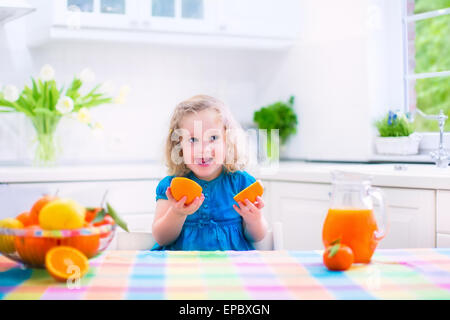 Cute funny little girl drinking freshly squeezed orange juice for healthy breakfast in a white kitchen with window Stock Photo