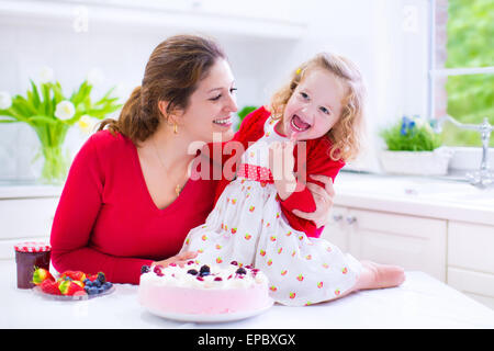 Happy family, young mother and her adorable little daughter, cute curly toddler girl in red dress, baking fresh strawberry cake Stock Photo