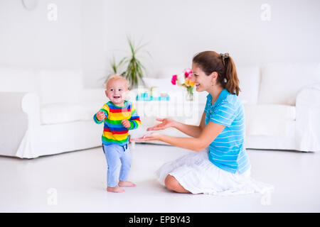 Cute little baby boy making his first steps, walking to his mother in a white sunny living room Stock Photo