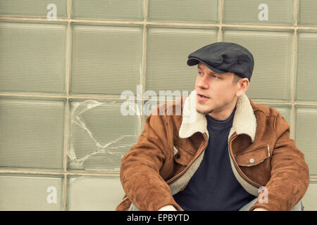 Cool guy in aviator jacket and newsie cap relaxes against a glass wall Stock Photo