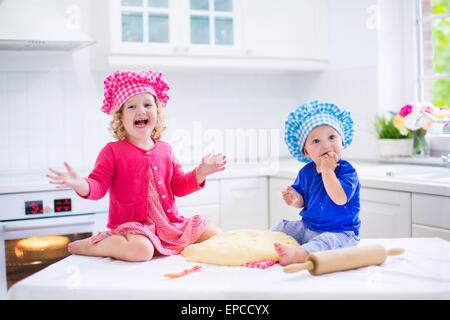 Cute kids, adorable little girl and funny baby boy wearing pink and blue chef hats playing with dough baking a pie in kitchen Stock Photo