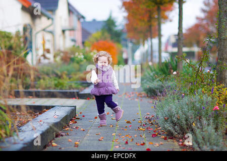 Little happy toddler girl in a warm purple jacket and boots playing in a beautiful city street with golden fall garden trees Stock Photo