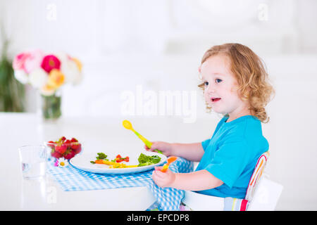Happy little girl, cute curly toddler, eating fresh vegetables for lunch, healthy salad snack, corn, broccoli, carrots Stock Photo