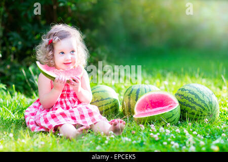 Funny little girl, adorable toddler with curly hair wearing a red dress, eating watermelon, healthy fruit snack Stock Photo