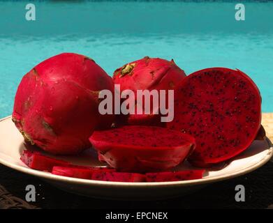 Whole, half, and pieces of red Dragon fruit on a white plate next to a swimming pool Stock Photo