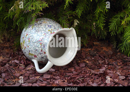 The antique jug with a pattern of flowers lies on brown chipped wood under a fir-tree. Stock Photo