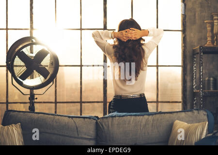Elegant brunette woman seen from behind, leaning against the back of a sofa in a loft. Hands laced behind her head, she is looking out the loft window. Industrial chic atmosphere. Stock Photo