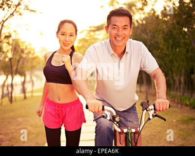 young asian couple running, riding bike outdoors in park at sunset, fitness, sport and exercise, healthy life and lifestyle conc Stock Photo