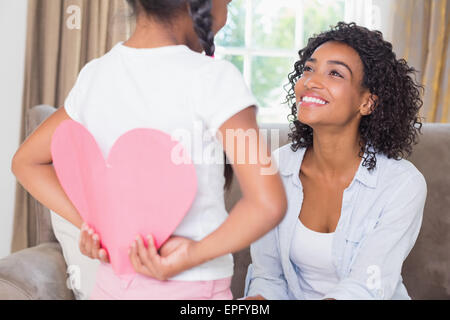 Pretty mother sitting on couch with daughter hiding heart card Stock Photo