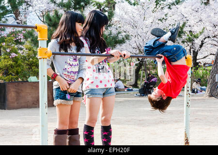 Two Japanese girls 10-11 year old, standing and watching Caucasian child, boy, 8-9 year old, swinging upside down on an exercise bar in park. Stock Photo