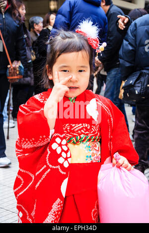 Very shy Japanese child, girl, 3-4 year old, wearing a red kimono and standing in front of viewer, giving the two finger peace gesture. Stock Photo