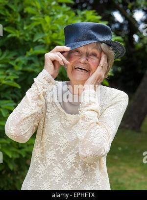 Portrait of a lively laughing elderly lady wearing a hat lifting her hand to the brim as she stands outdoors in a lush green gar Stock Photo
