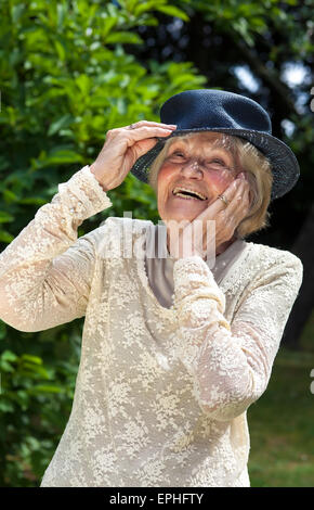 Vivacious elderly lady wearing a hat and elegant lace top laughing outdoors in a summer garden. Stock Photo
