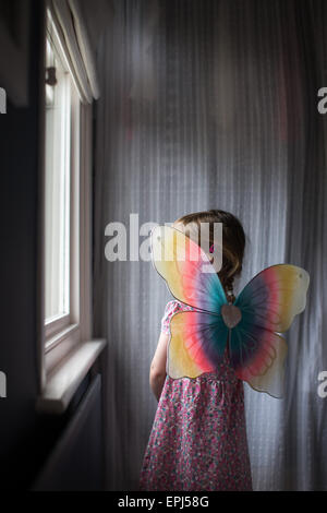 A child aged 3 looks out of a bedroom window wearing brightly coloured fairy wings Stock Photo