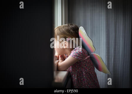 A child aged 3 looks out of a bedroom window wearing brightly coloured fairy wings Stock Photo