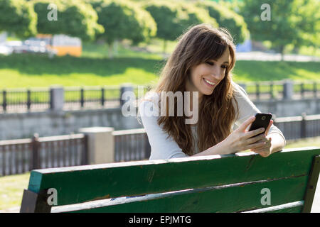 Young beauty woman have fun with using smartphone sitting on bench. Happy caucasian girl texting with phone in city park Stock Photo