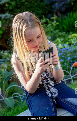 Blonde 8 year old girl sitting in a garden looking at a mobile phone Stock Photo