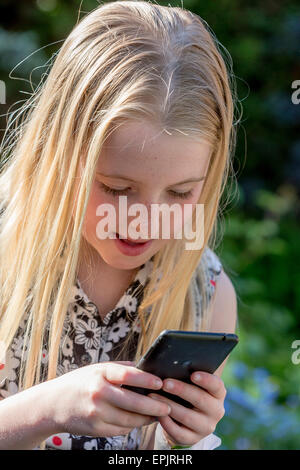 Blonde 8 year old girl sitting in a garden looking at a mobile phone Stock Photo