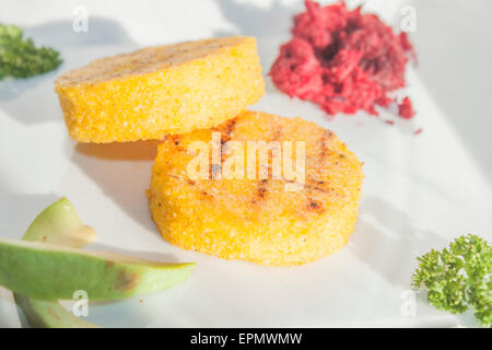 Two yellow polenta slices, near avocado, parsley and beetroot, on a white table Stock Photo