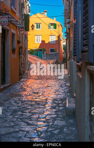 Morning sun shines on an orange building in Rovinj, Croatia, and casts an orange glow brightly on the wet cobble stones below. Stock Photo