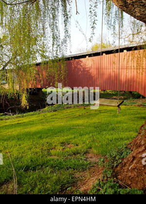 A tree swing hangs from a willow tree near a red covered bridge over a stream in the springtime. Stock Photo