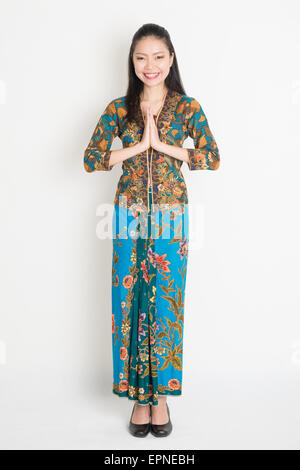 Full length Southeast Asian woman with batik dress in greeting gesture standing on plain background. Stock Photo