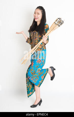 Full body portrait of happy Southeast Asian woman in batik dress hand holding bamboo oil lamp standing on plain background.