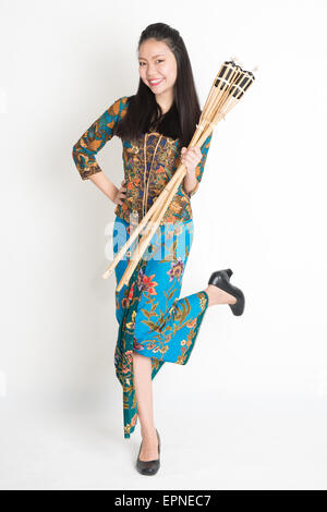 Full body portrait of Southeast Asian woman in batik dress hands holding bamboo oil lamp standing on plain background.