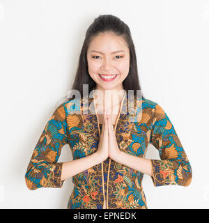 Portrait of happy Southeast Asian woman with batik dress in greeting gesture on plain background. Stock Photo