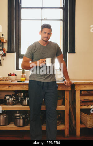 Portrait of young man standing in kitchen holding a cup of coffee.  Handsome male model having fresh morning coffee at home in k Stock Photo