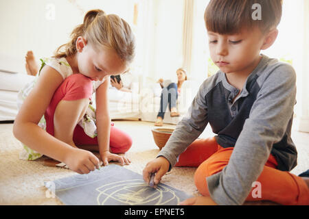 Little siblings drawing a picture with chalk colors. Cute children sitting on floor with parents in background in living room at Stock Photo