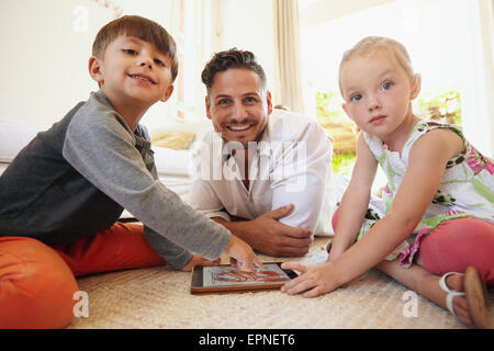 Family sitting on floor using digital tablet looking at camera smiling. Father with son and daughter playing with tablet compute Stock Photo