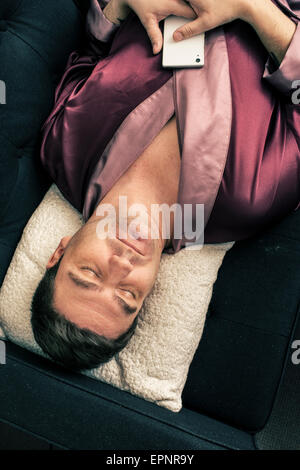 Man lying and resting on sofa at home. He is sleeping with his head on a pillow and holding a cell phone in his hands. Stock Photo