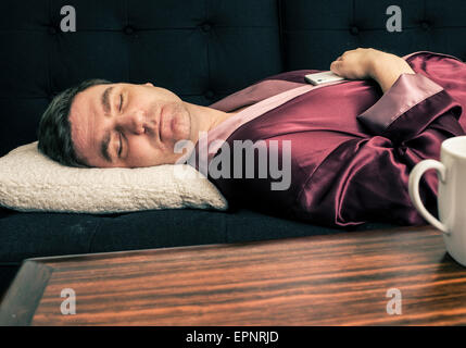 Man lying and resting on sofa at home. He is sleeping with his head on a pillow and holding a cell phone in his hands. Stock Photo