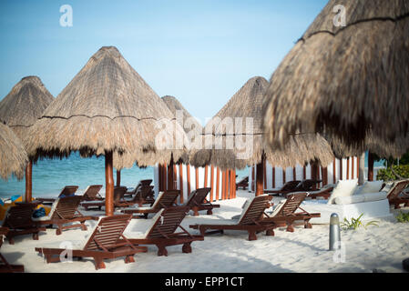 Beach Deck Chairs Playa Mujeres Mexico | PLAYA MUJERES, Mexico — Rows of palapa-shaded loungers line the white sand at the Beloved Hotel on Playa Mujeres, a beachfront resort in the Costa Mujeres corridor north of Cancun. The palm-thatched umbrellas are a standard feature of Mexican Caribbean resorts, providing shade against the intense midday sun typical of the Yucatan Peninsula's tropical climate. Stock Photo