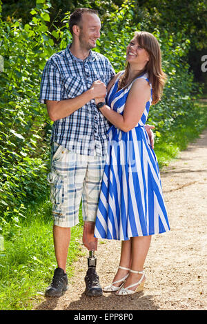 Happy attractive couple in love standing in a close embrace on a leafy green pathway with the man wearing a prosthetic leg. Stock Photo