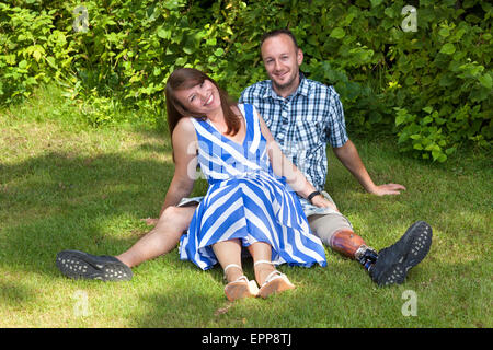 Happy attractive couple relaxing in the garden sitting joyful close together on the grass - man with leg prosthesis. Stock Photo