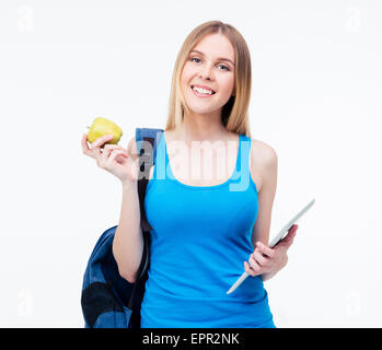 Smiling casual woman with backpack holding green apple and tablet computer. Standing isolated on a white background. Looking at  Stock Photo