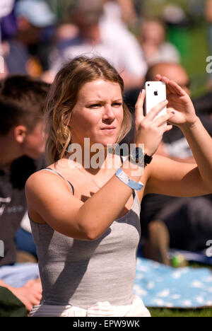 Young female amongst the crowds taking photos on her iPhone of local bands at a music festival (CHIDDFEST 2015), Surrey, UK. Stock Photo