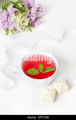 Cup of hibiscus tea with mint leaves, white chocolate candies and flower bouquet on the white table at breakfast time Stock Photo
