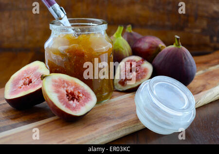 Fresh figs on wooden cutting chopping board with jar of fig jelly preserve on dark wood rustic table background. Stock Photo