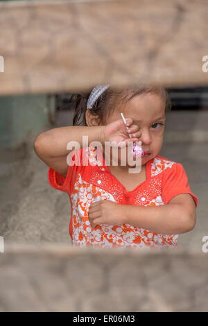 Little girl with candy sucker wiping her eyes after crying as viewed through a fence in Guatemala Stock Photo