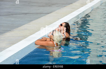 Two women, senior blonde mother and mid adult brunette daughter are standing in bright blue water at the edge of swimming pool a Stock Photo