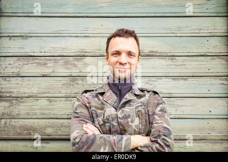 Young smiling Caucasian man in camouflage, outdoor portrait over green rural wooden wall Stock Photo
