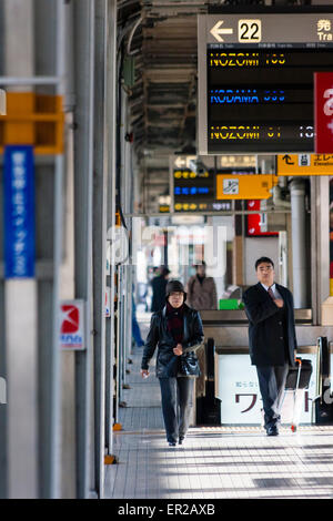 Compressed perspective view along platform 22 of Shin Osaka station with two people walking towards viewer and various over head information signs. Stock Photo