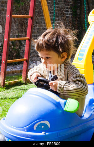 Young child, boy, 3-5 year old driving in play peddle car in a garden, with a concentrated look on his face as both hands grip the steering wheel. Stock Photo