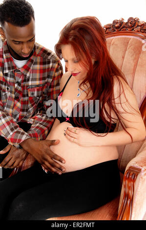 A red hair young pregnant women sitting in a pink armchair and her husband hold his hand on her belly, isolated for white Stock Photo