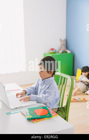 Elementary age boy doing his homework at his desk with his sister reading on the floor in the background Stock Photo